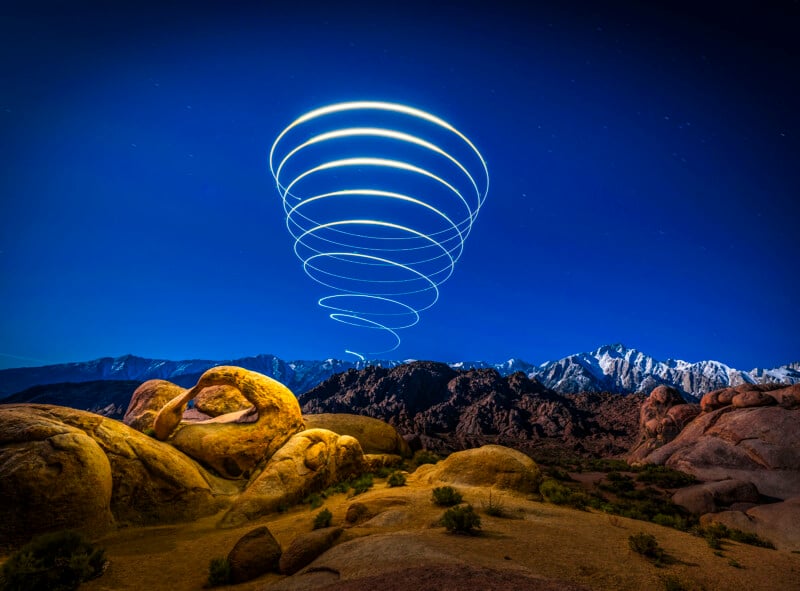 A rocky desert landscape under a deep blue night sky, with mountains in the background. Glowing spiral light trails rise above the rocks, creating a mesmerizing pattern above the horizon.