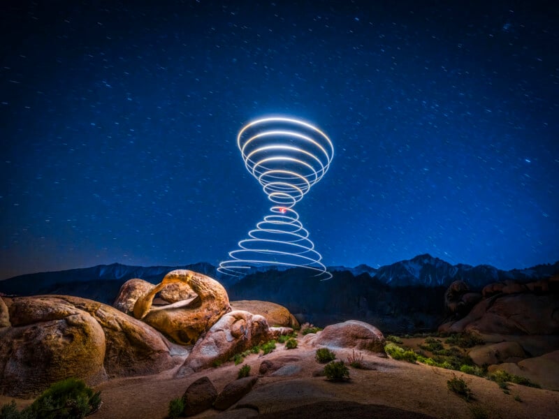 A night sky over rocky terrain shows spiraling light trails creating a tornado-like shape, with stars scattered across the deep blue sky and mountains in the background.