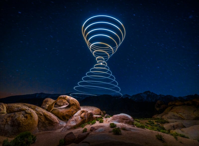 Rocks and shrubs under a night sky with distant mountains; spiral patterns of light trail upward in the air, creating a glowing vortex effect above the landscape.