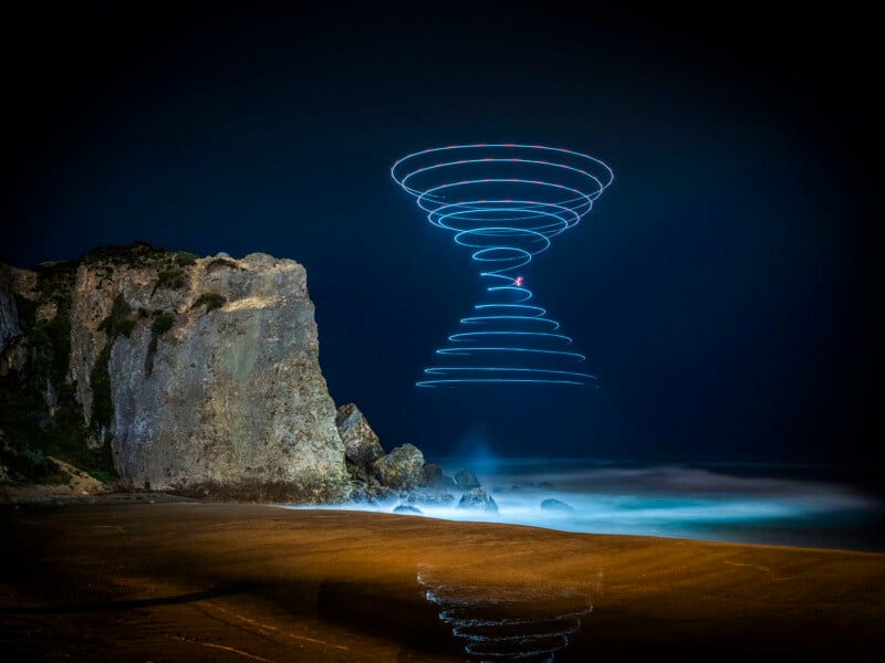 A rocky beach at night with a spiral pattern of blue and red light drawn in the sky, likely from a long-exposure photo of a drone, reflected on the wet sand below.