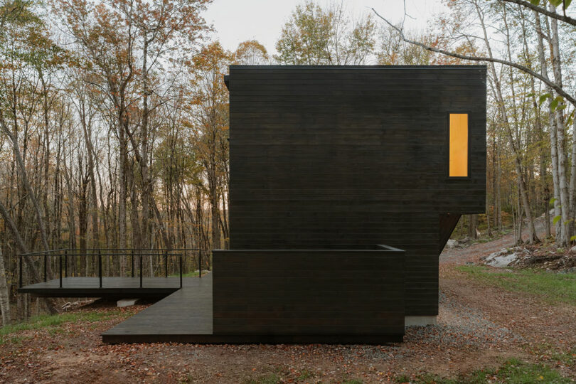 A modern, dark-wooden rectangular cabin with large decks sits in a forested area; one window is lit, and autumn trees surround the structure.