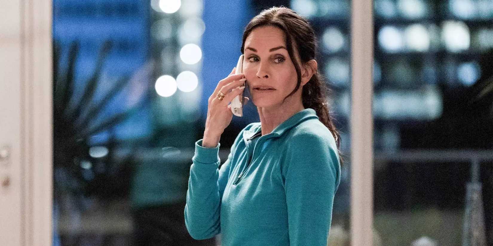 Gale Weathers (Courteney Cox), in a blue hoodie, talks on the phone while looking over her shoulder in Scream VI