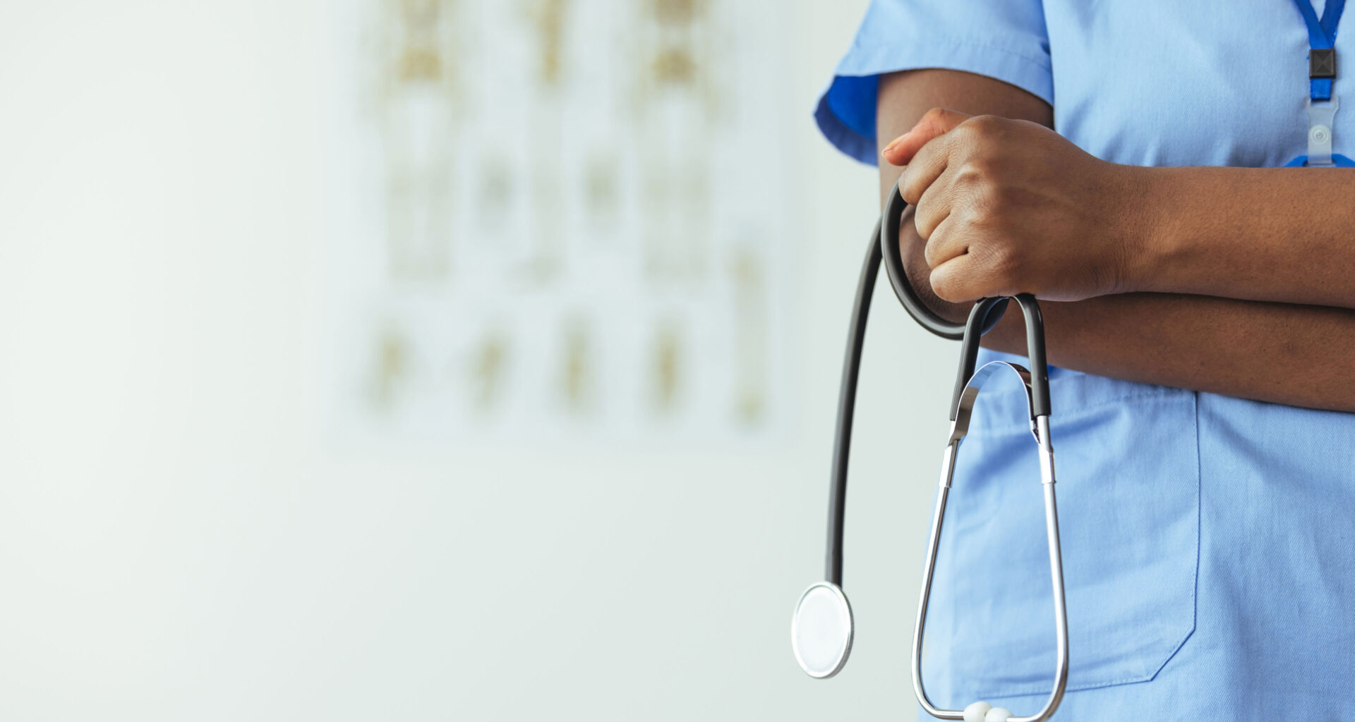 An unidentifiable medical professional stands to the side of frame in blue scrubs and holds a stethoscope.