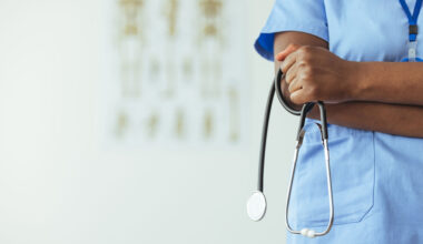 An unidentifiable medical professional stands to the side of frame in blue scrubs and holds a stethoscope.