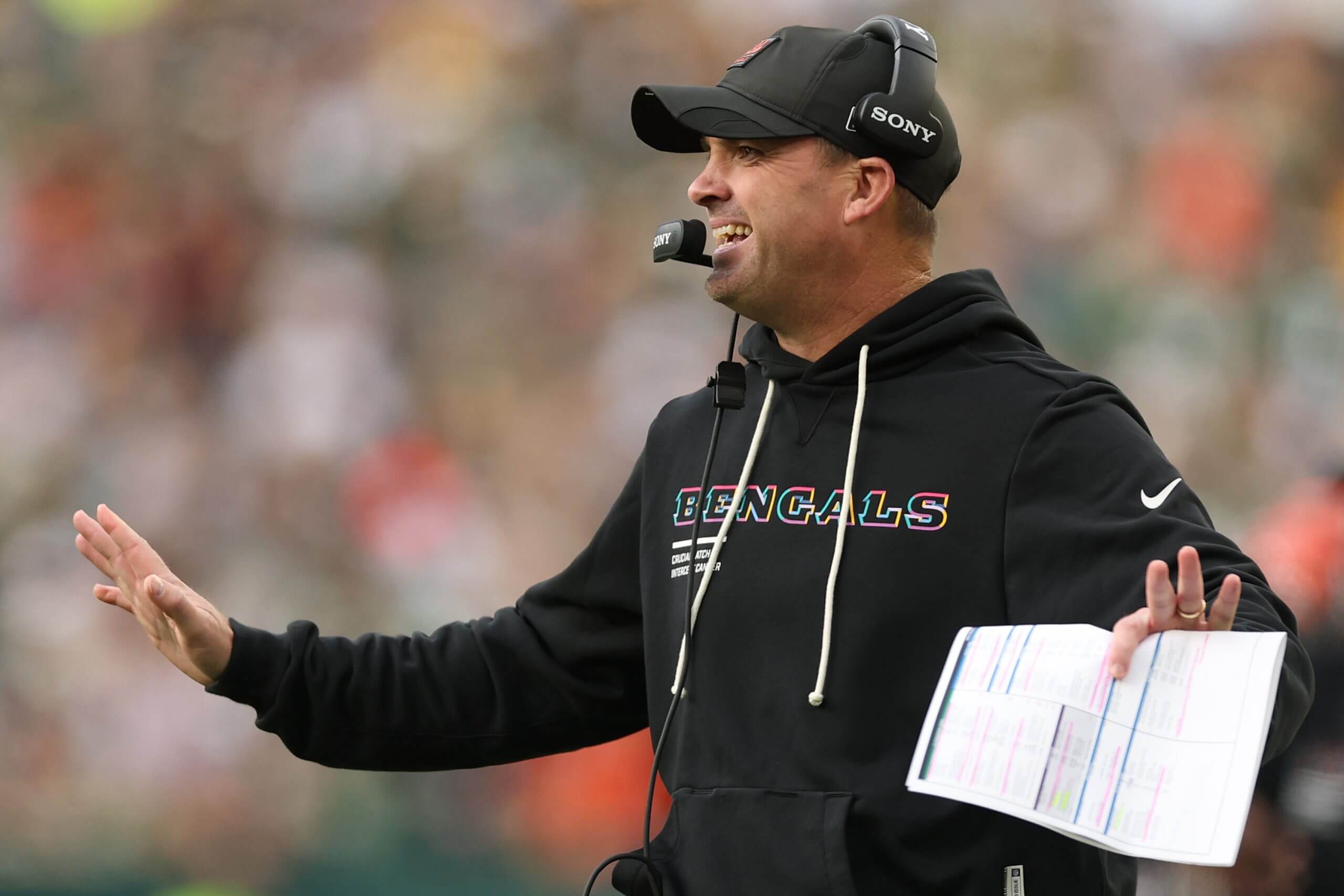 Zac Taylor, wearing a black hoodie and hat with a headset, smiles while holding his arms out to his sides, with a play sheet in his left hand.
