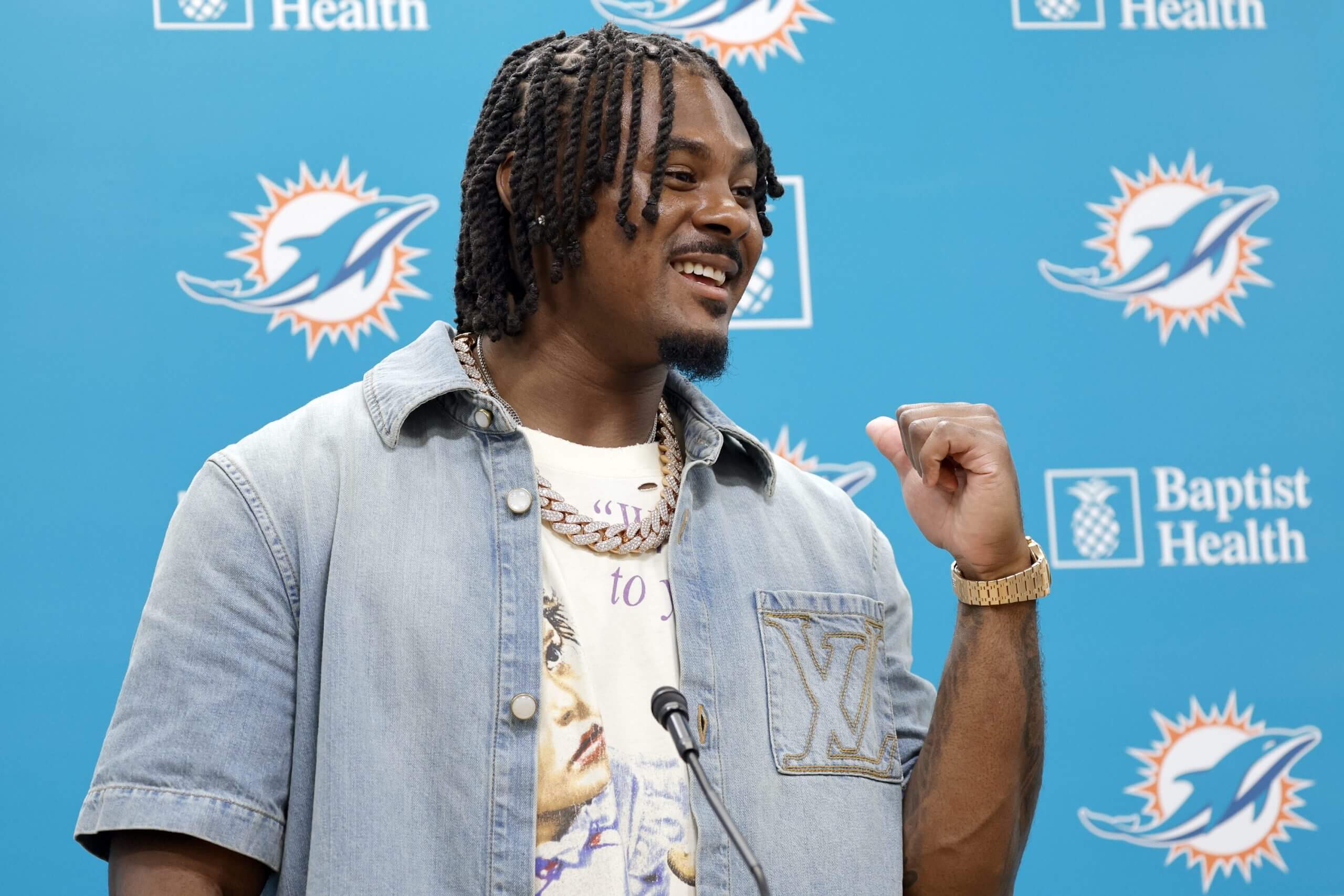 Malik Willis, wearing a denim shirt, smiles while standing at a microphone in front of an aqua backdrop with the Dolphins' logo.