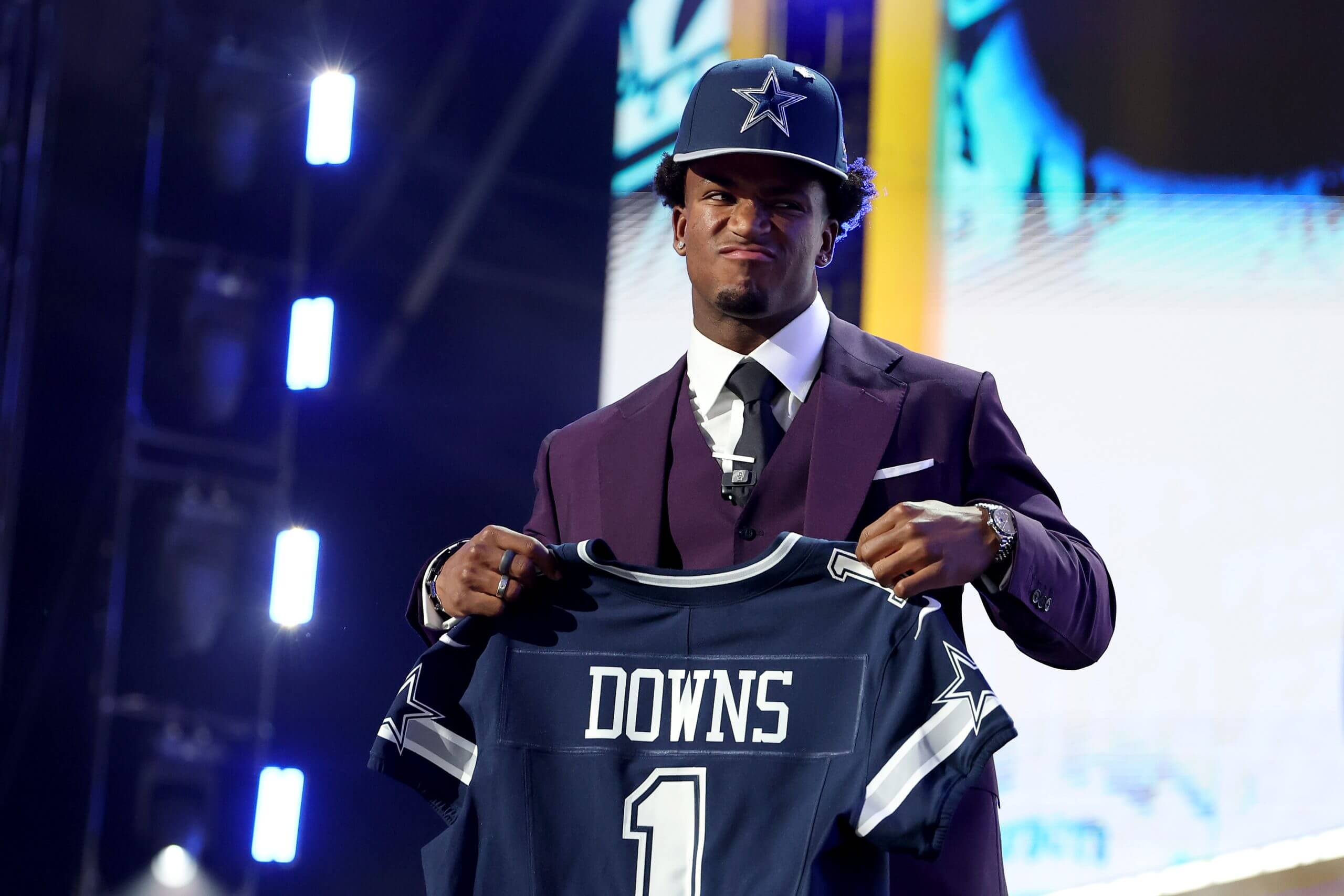 Caleb Downs, wearing a purple suit and a navy blue Cowboys hat, holds up a Cowboys jersey and looks to his left.