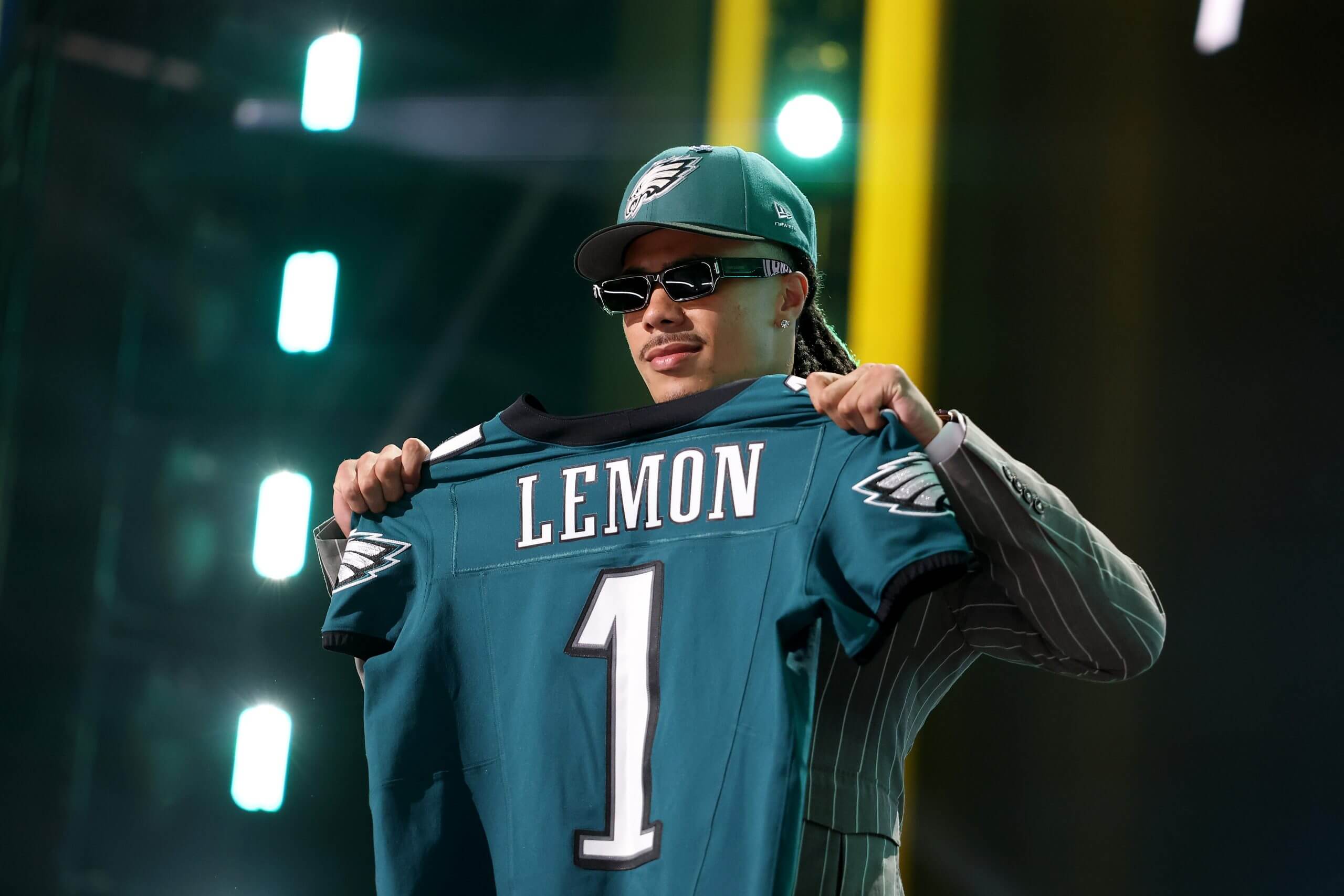 Makai Lemon, wearing a striped suit, sunglasses and a green Eagles hat, holds up an Eagles uniform.