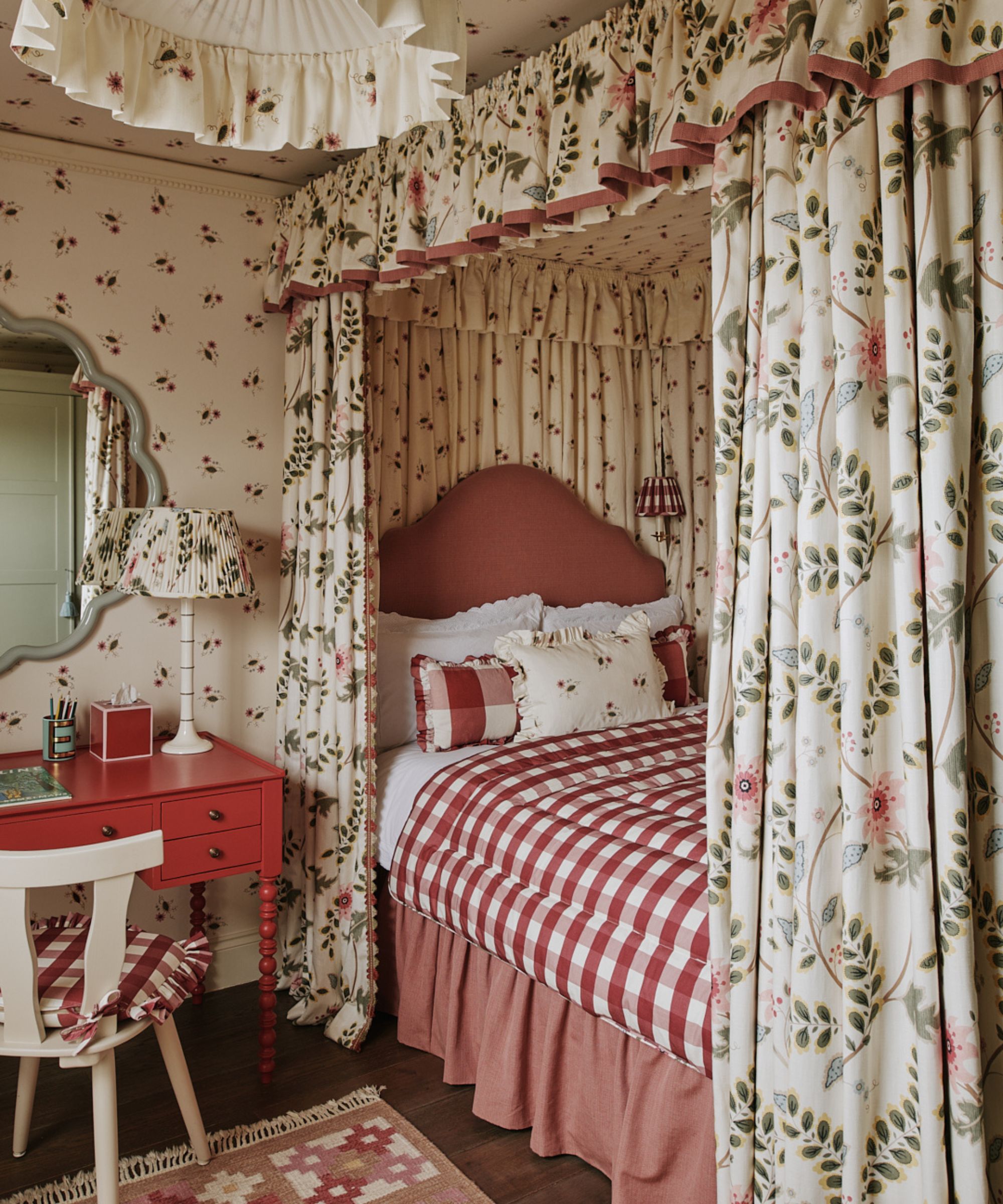 A cozy pattern-filled bedroom with a canopy bed with cream, pink, and green floral-patterned upholstery, red and white gingham bedding, a dusky pink bed skirt, wallpapered walls, and a red vanity.