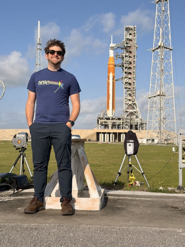A man wearing sunglasses and a NASA t-shirt stands smiling in front of NASA's Artemis I Space Launch System rocket on its launch pad, with blue sky and clouds in the background.