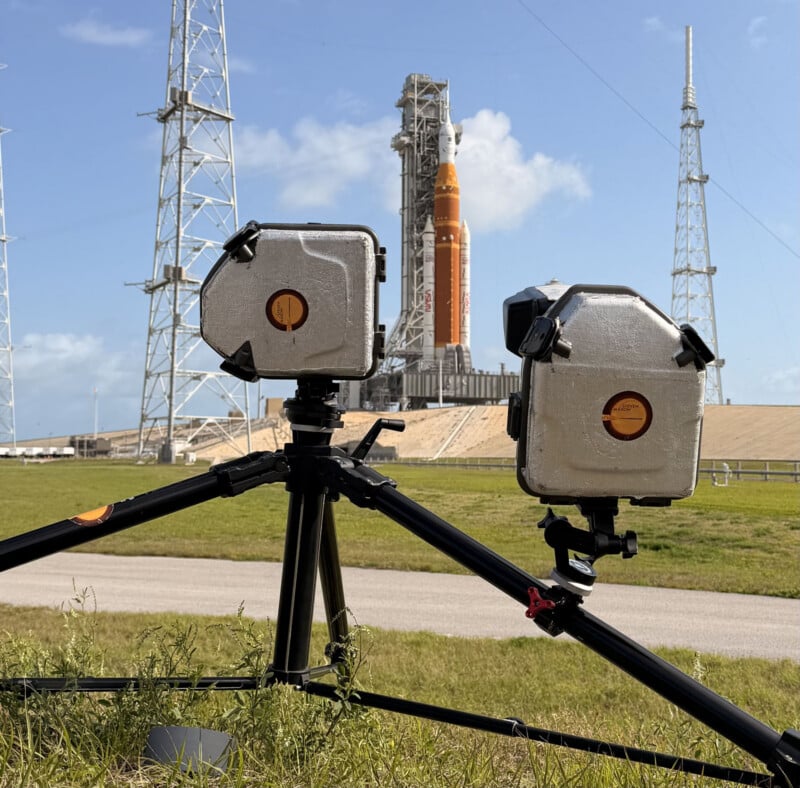 Two weatherproof cameras on tripods are set up on a grassy area, aimed at a large orange and white rocket on a launch pad in the background, with blue sky and metal towers visible.