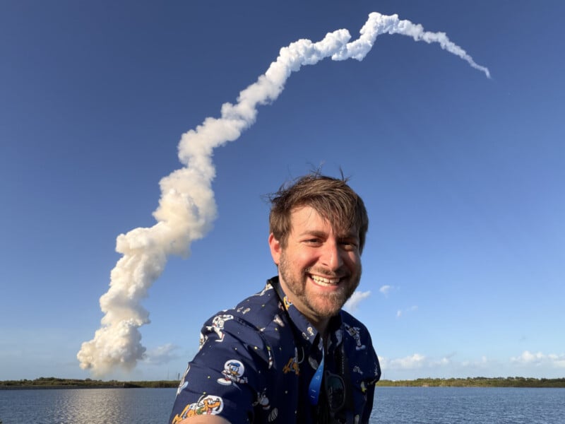 A smiling man in a space-themed shirt stands near water, with a rocket launch visible behind him, leaving a curving trail of smoke in the clear blue sky.