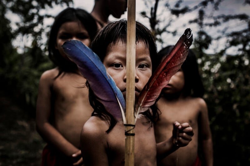 A young child holds a stick decorated with two colorful feathers, one blue and one red, in front of their face while three other children stand barefoot in the background outdoors. They all have dark hair and are shirtless.
