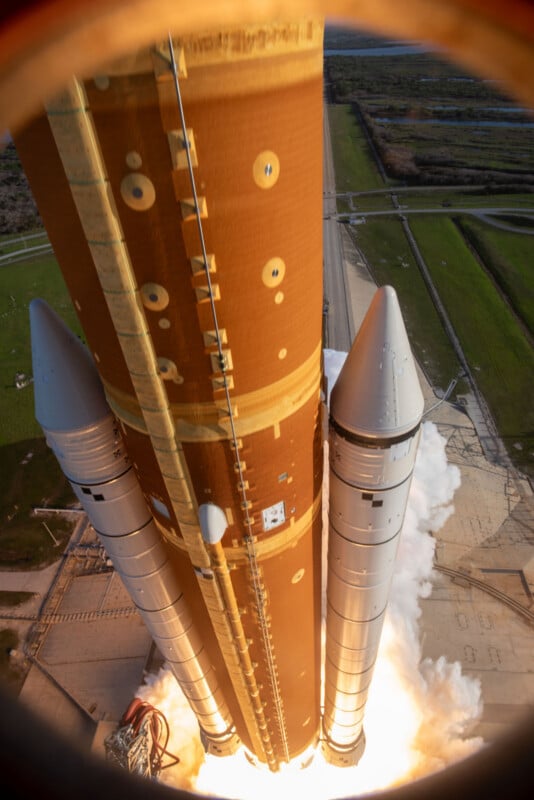 A rocket launches vertically, viewed from above with engines igniting and smoke billowing below. The image shows the rocket’s main fuel tank and two solid rocket boosters, with green fields and a runway in the background.