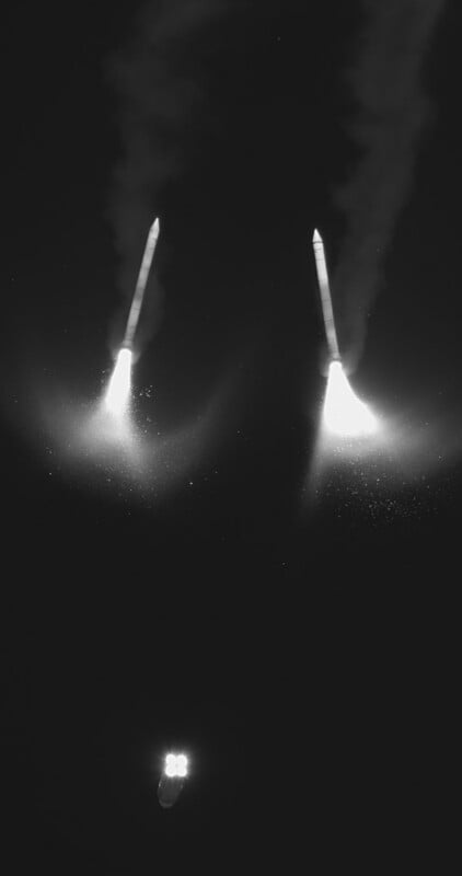 A black-and-white image shows three rocket boosters descending through the night sky, each with bright flames and visible trails of smoke beneath them. Two boosters are near each other at the top, and one is lower down.