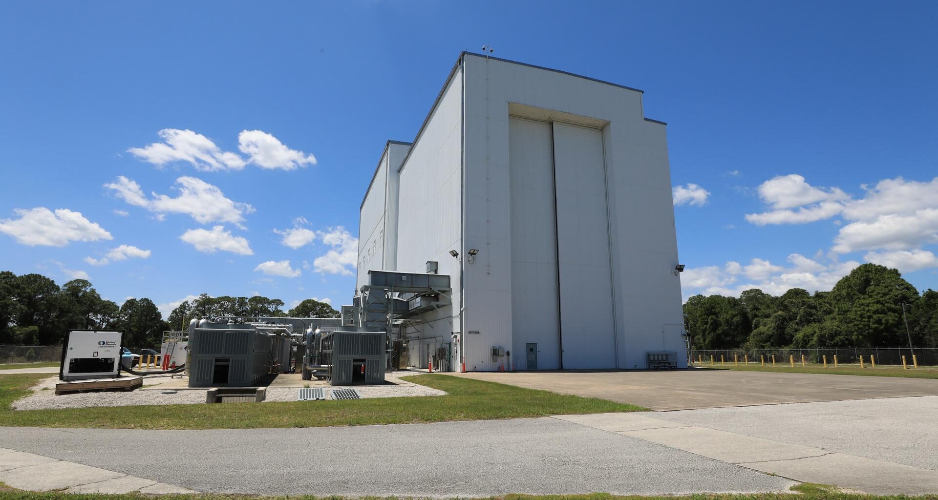 Image shows sunny, blue skies with a few wispy clouds outside the NASA Kennedy’s Payload Hazardous Servicing Facility (PHSF). The building is a grayish color with a nearby driveway and a small door for people to enter and huge doors for spacecrafts and instruments to enter. Photo credit: NASA/Kim Shiflett