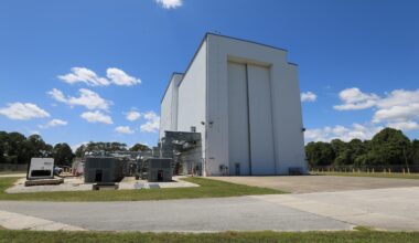 Image shows sunny, blue skies with a few wispy clouds outside the NASA Kennedy’s Payload Hazardous Servicing Facility (PHSF). The building is a grayish color with a nearby driveway and a small door for people to enter and huge doors for spacecrafts and instruments to enter. Photo credit: NASA/Kim Shiflett