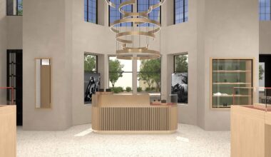 Modern retail store interior with a light wood counter in the center, minimalist shelves, large windows letting in natural light, and a circular, layered chandelier hanging from the ceiling. Trees are visible outside.