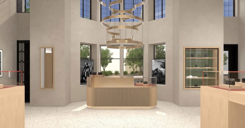 Modern retail store interior with a light wood counter in the center, minimalist shelves, large windows letting in natural light, and a circular, layered chandelier hanging from the ceiling. Trees are visible outside.