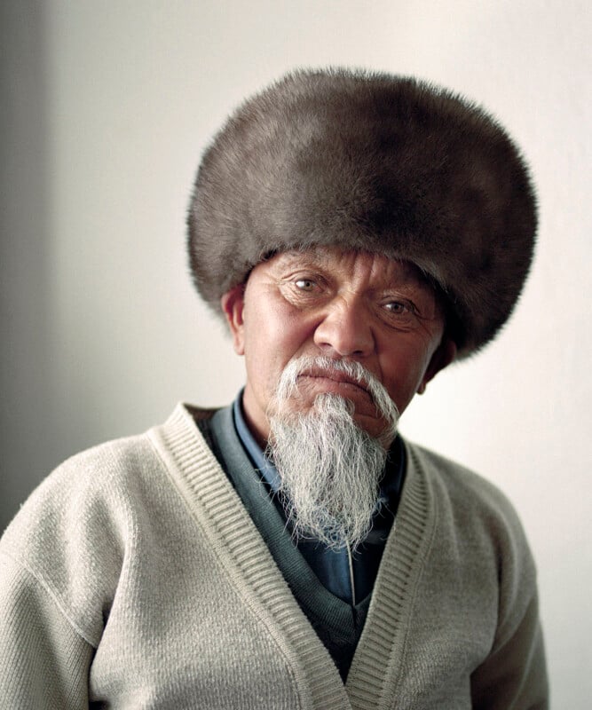 An older man with a long white beard wears a large, round fur hat and a beige cardigan over a collared shirt. He is looking at the camera with a neutral expression, against a plain light background.