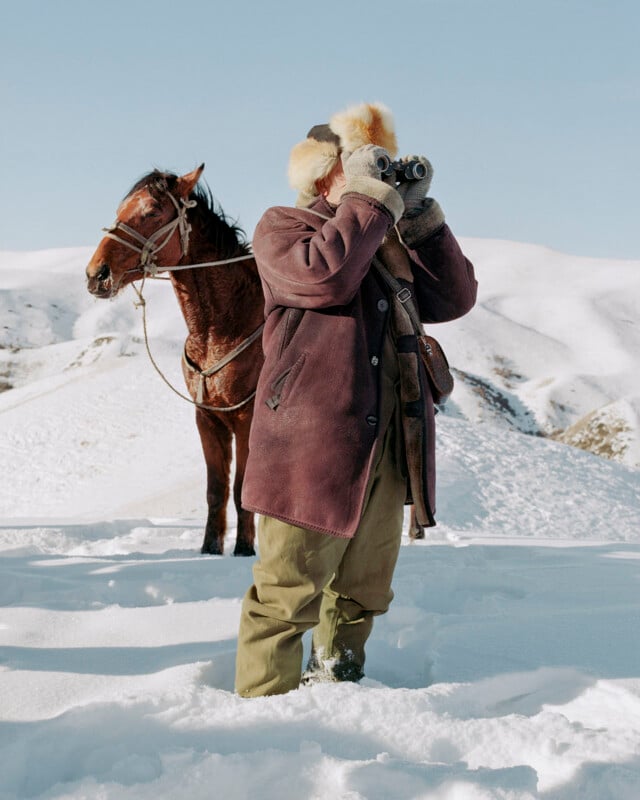 A person wearing a fur hat and heavy winter coat stands in deep snow, looking through binoculars. A saddled brown horse stands behind them against a backdrop of snowy hills under a clear sky.