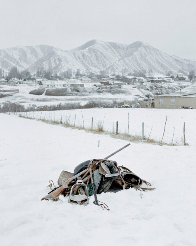 A pile of old, rusty farm tools and metal objects sits in a snow-covered field, with a snowy mountain range and scattered houses in the background under an overcast sky.