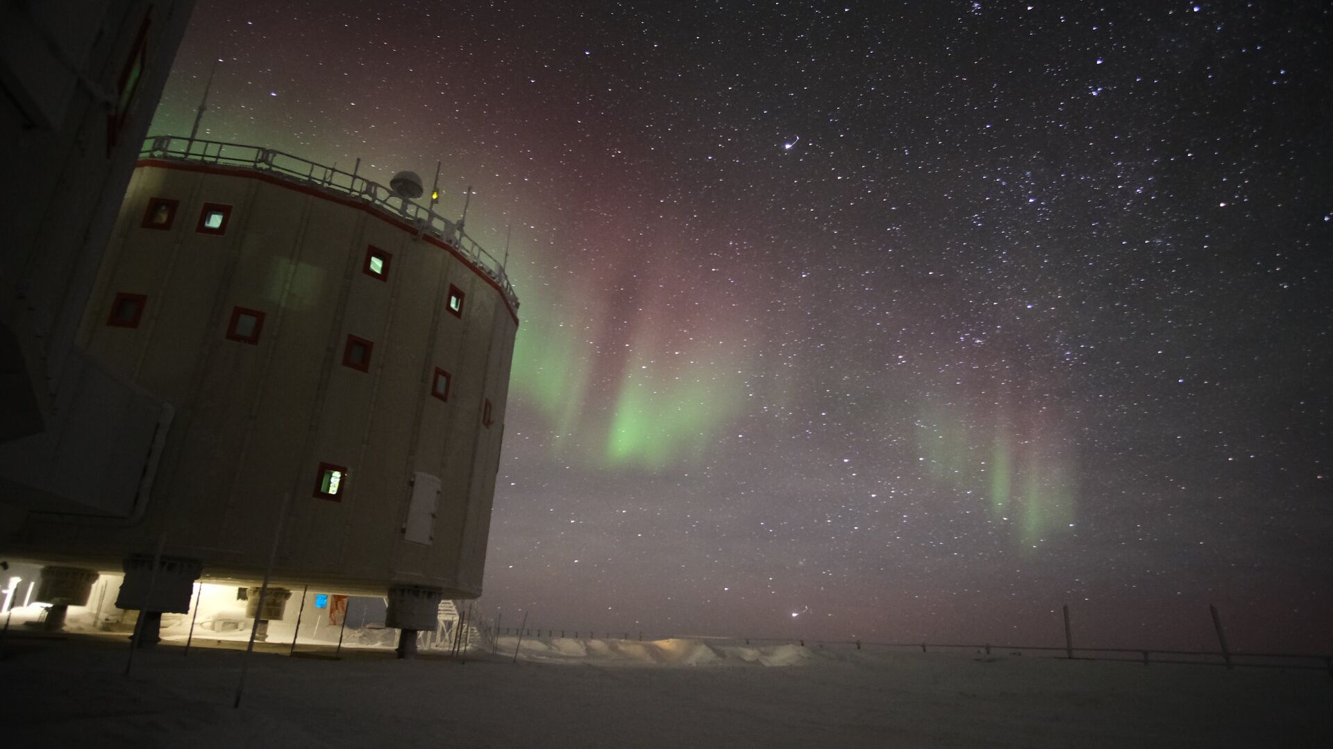 cylindrical building with lots of little windows. the southern lights dance in the sky behind, ribbons of green and magenta against a sky full of stars.