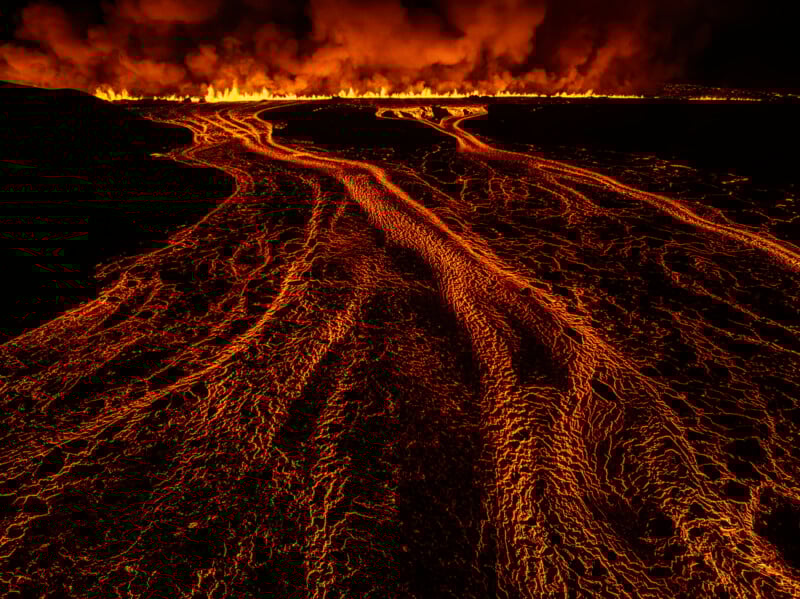 Molten lava flows down a dark landscape, glowing bright orange and red. In the background, a line of fire burns intensely, with thick smoke billowing upwards into the night sky.
