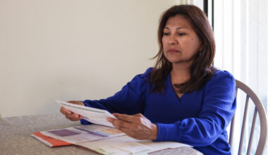 A Hispanic woman sits at a table, reading medical bills from a red file folder in front of her.