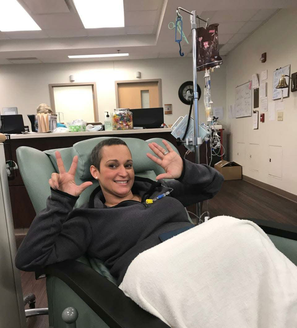 Marielle Santos McLeod poses, smiling, during chemo treatment. She holds up fingers on her left and right hands, totaling eight.