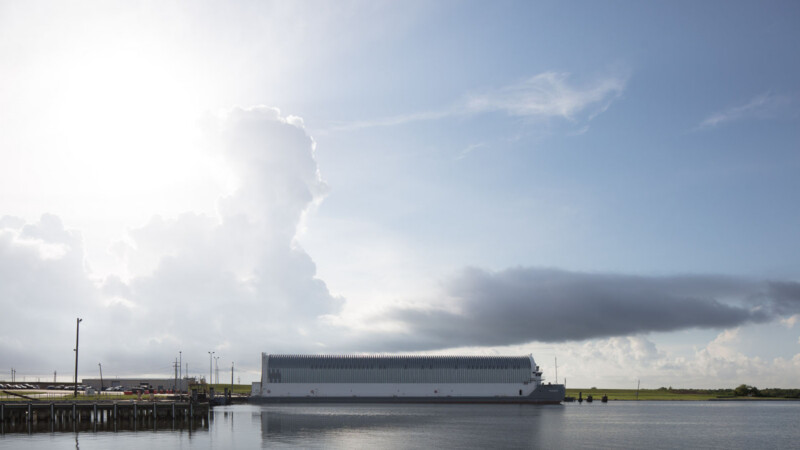 A large, rectangular white building floats on calm water near a shoreline, under a bright sky with dramatic clouds and sunlight streaming in from the left. The scene is peaceful and expansive.