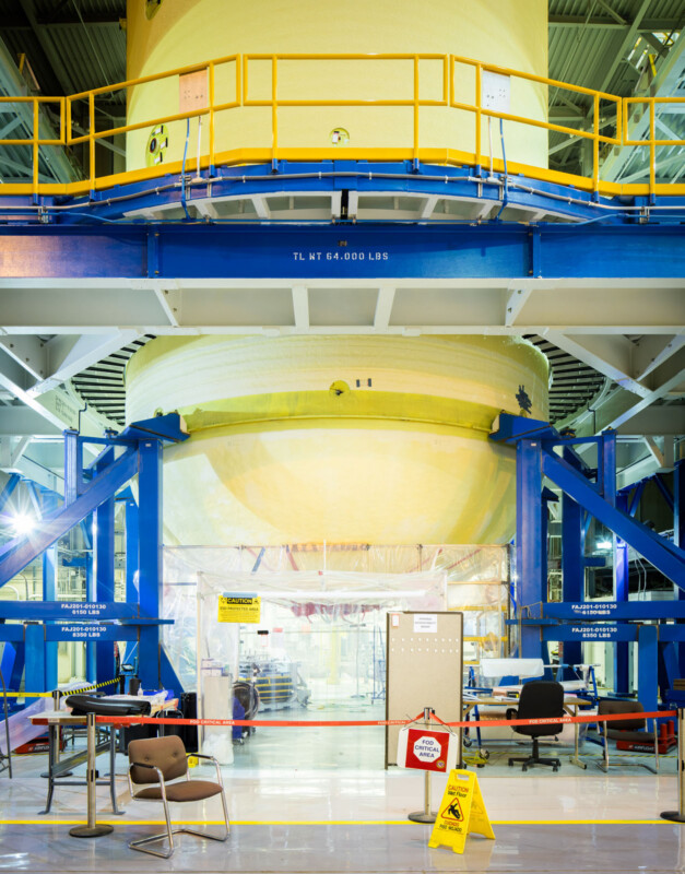 A large yellow cylindrical rocket component is suspended above the ground inside an industrial facility, supported by blue structures. Chairs, desks, and a caution sign are visible in the foreground.