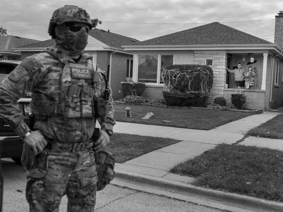 A black-and-white image shows a heavily armed police officer standing in a suburban street while two residents watch from a house decorated with Halloween ornaments.