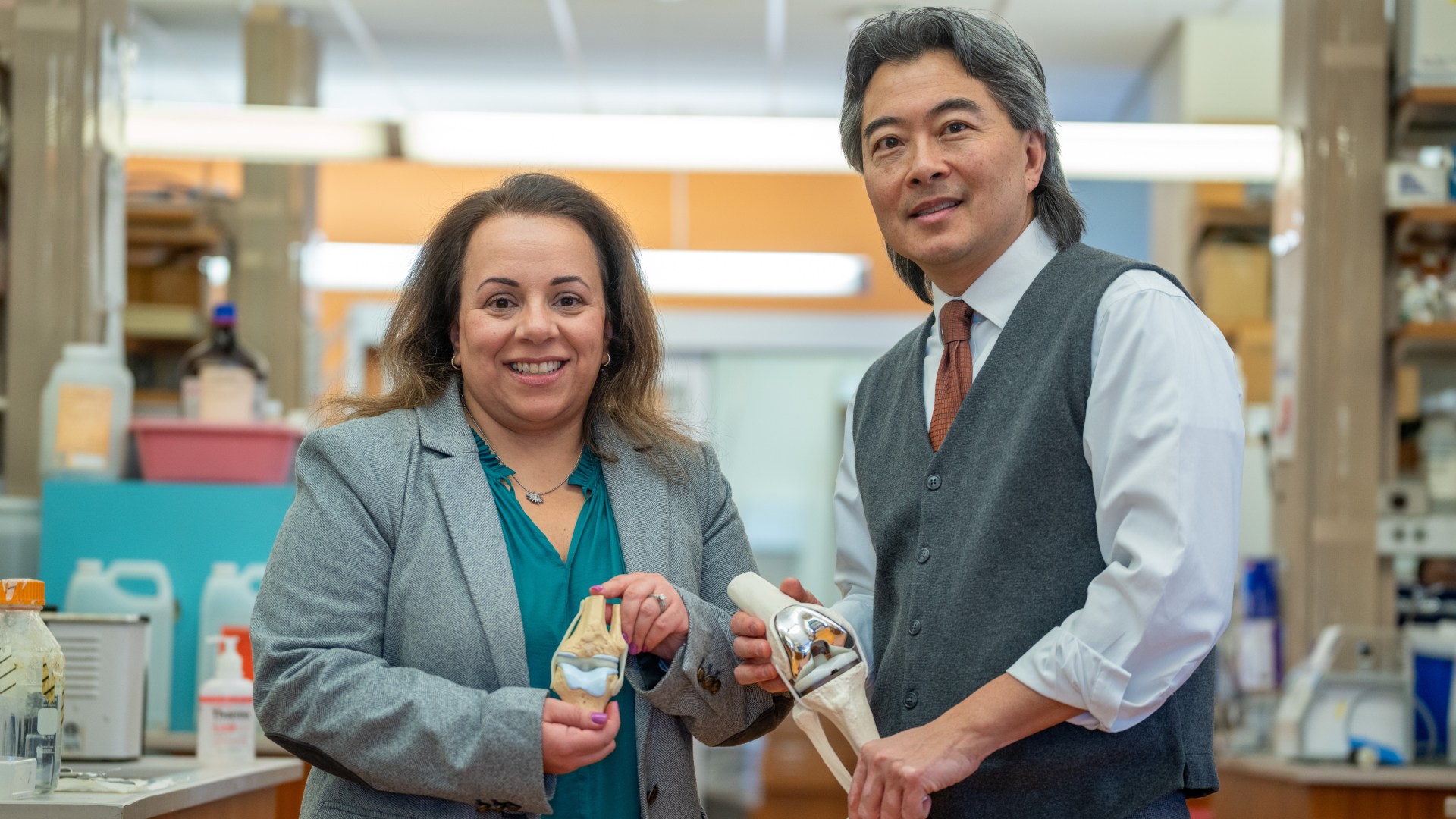 A man with long gray hair wearing a brown vest and white shirt speaks with a woman with long brown hair as they both look at various prosthetic limbs.