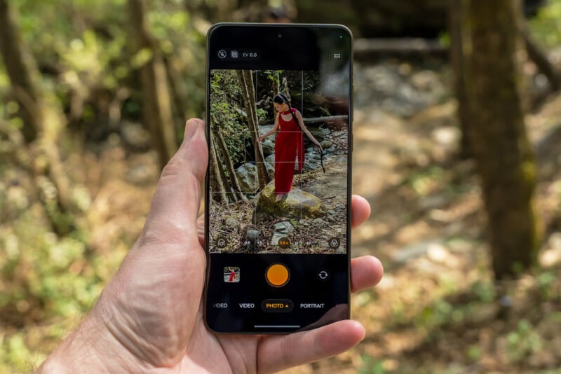 A hand holds a smartphone outdoors, displaying its camera app. The screen shows a woman in a red dress standing on a rock in a wooded area, matching the blurred natural background behind the phone.