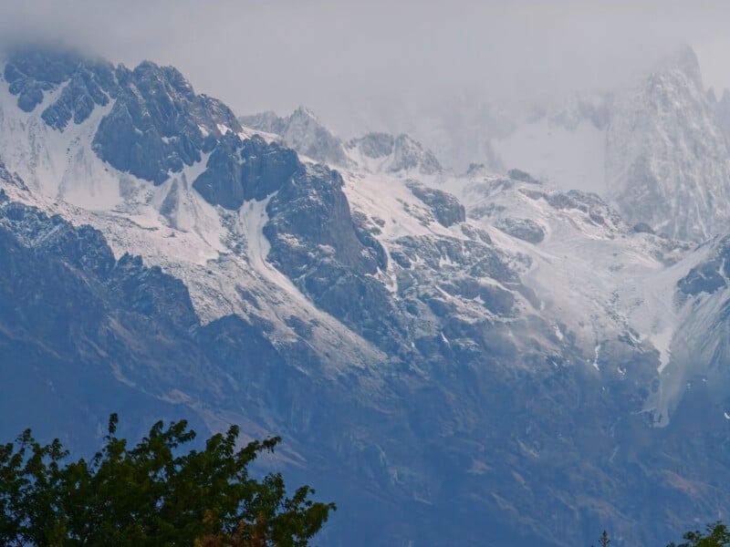 Snow-covered rocky mountain peaks under a cloudy sky, with green tree branches in the foreground.