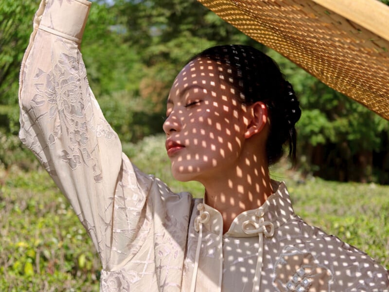 A woman in a white embroidered outfit stands outdoors, eyes closed, holding a woven hat above her head. Dappled sunlight creates a grid-like pattern of light and shadow on her face and clothes. Green foliage is in the background.