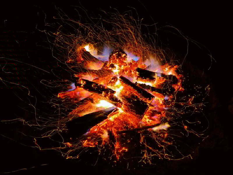 A campfire burns brightly at night, with orange flames, glowing embers, and flying sparks surrounding the stacked logs against a dark background.