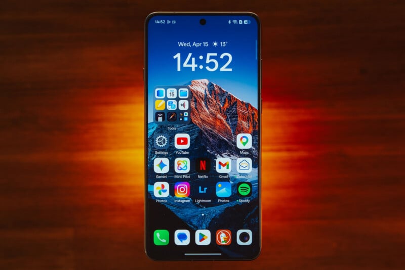 A smartphone with a mountain wallpaper displays the time 14:52 and several app icons, including YouTube, Gmail, Instagram, and Spotify, against a wooden surface with an orange light glow behind the phone.