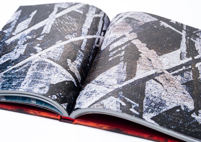 An open book displaying abstract art with intersecting white and gray lines on a dark, textured background. The book's spine and edge are visible against a white surface.