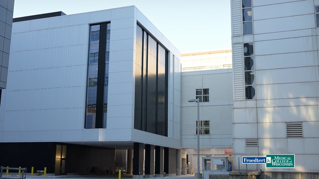 Modern medical building with large dark windows and white exterior panels, featuring a sign for Froedtert & Medical College of Wisconsin in the lower right corner.