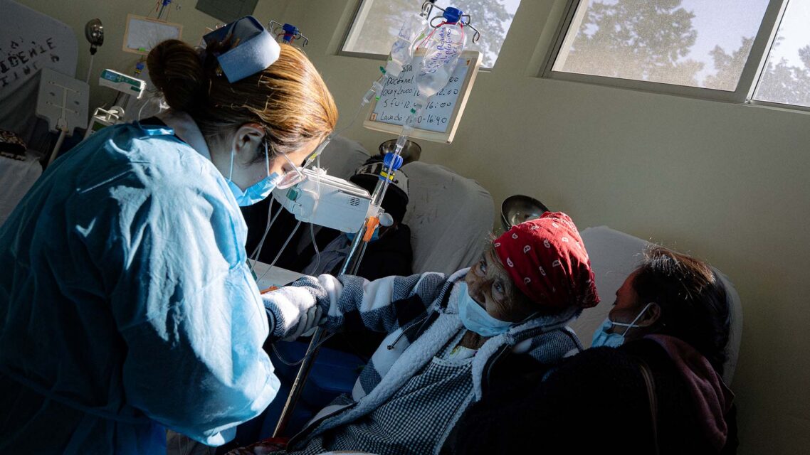 Photo of a nurse in a protective gown assisting a patient who has an IV bag.