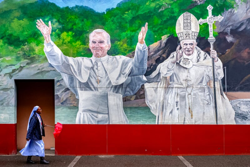A nun in a blue habit walks past a colorful mural depicting two popes in white robes and ornate headpieces, with one smiling and raising his arms, set against a backdrop of greenery and rocks.