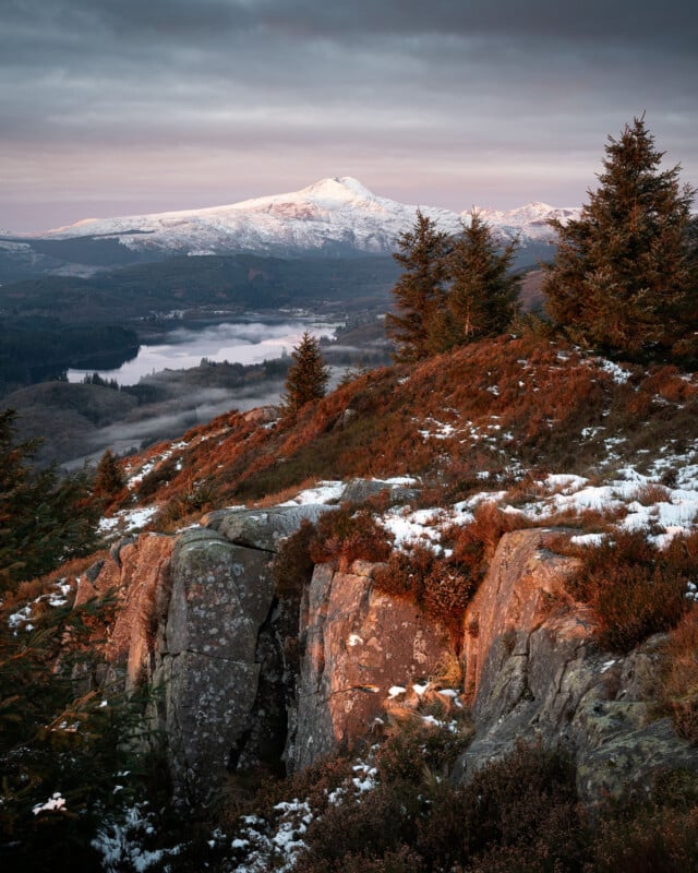 A scenic mountain landscape at sunrise, with snow-capped peaks in the background, evergreen trees, rocky cliffs, and patches of snow on the foreground hillside bathed in warm sunlight. A lake and forest are visible below.