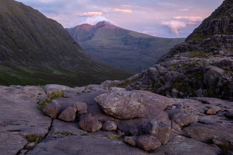 Rocky foreground with large stones, leading to a green valley between steep mountains; a distant sunlit peak is visible under a partly cloudy, purple-tinged sky at sunset.