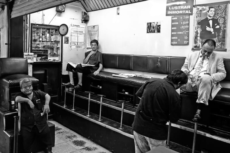 Black and white photo of a shoe-shine shop with three men sitting on raised chairs and two workers below; one worker leans on a railing, while the other sits and reads a newspaper. Price list and clock are visible on the wall.