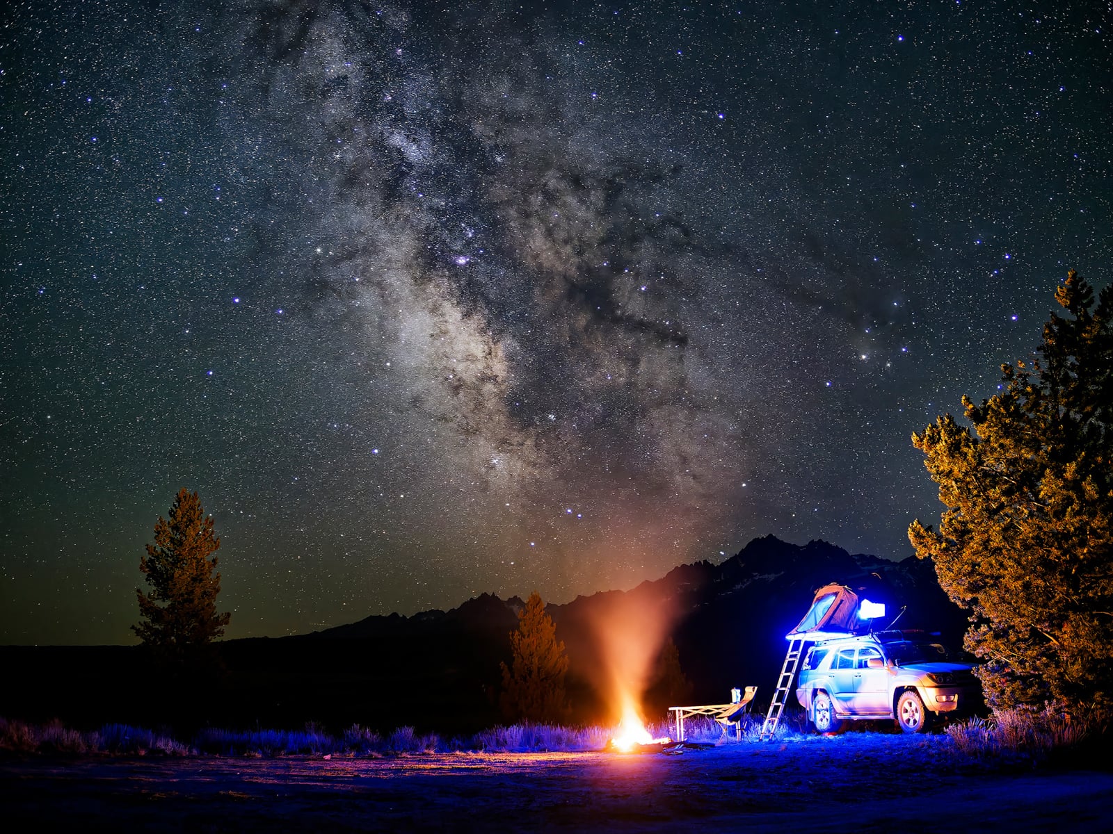 A car with a rooftop tent is parked under a star-filled night sky with the Milky Way visible. A campfire burns nearby, casting light on camping chairs and surrounding trees.
