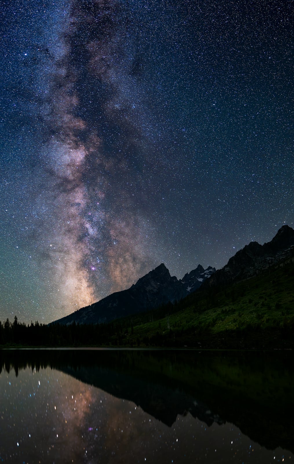 The Milky Way stretches across a star-filled night sky above silhouetted mountains and a grassy landscape, with the scene reflected in a calm, dark lake below.