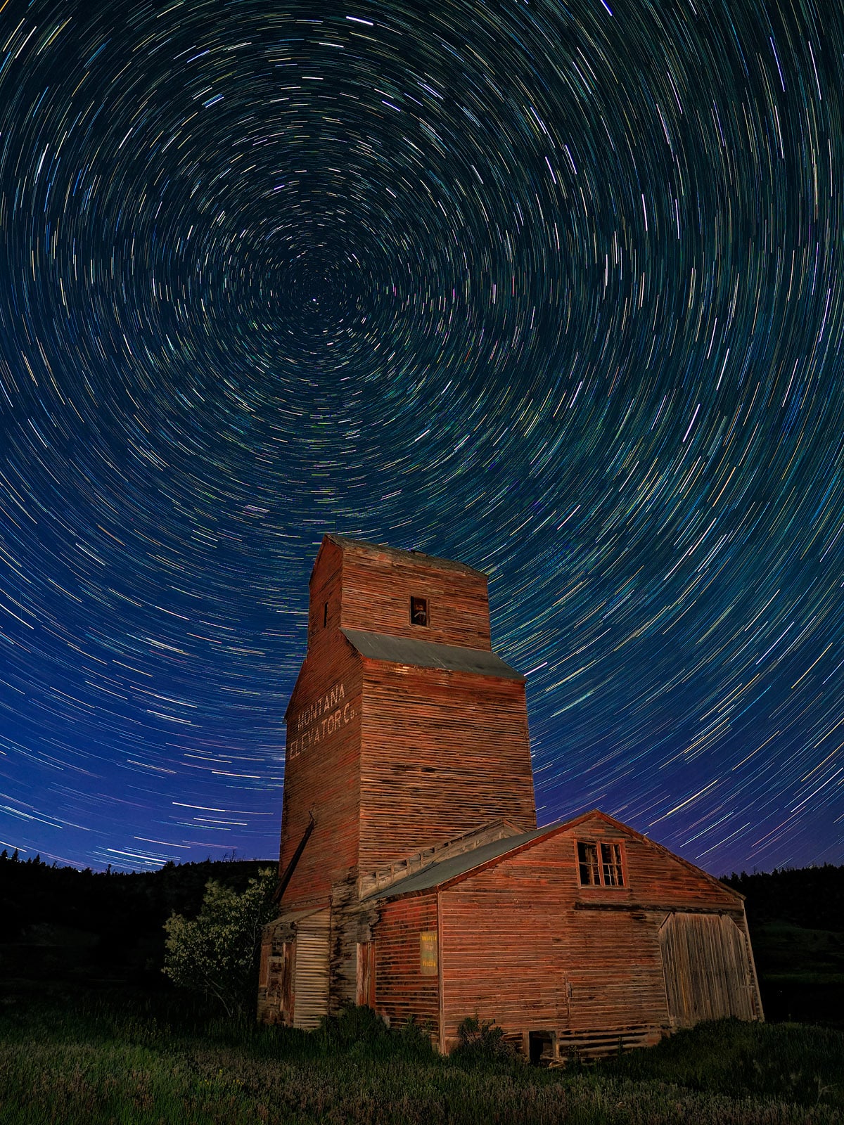 An old wooden grain elevator stands under a night sky filled with swirling star trails, creating circular patterns above the building, which is illuminated from below.
