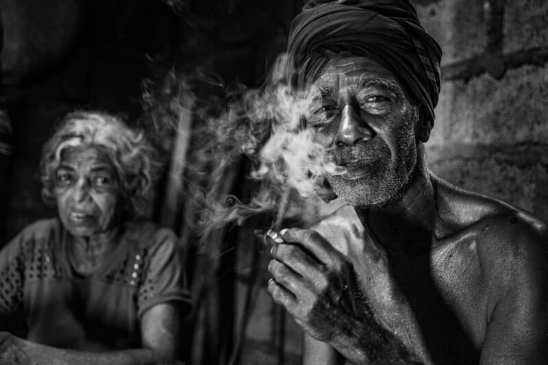 An elderly man wearing a turban smokes while looking at the camera, with smoke swirling around his face. An elderly woman sits slightly blurred in the background. The photo is in black and white.